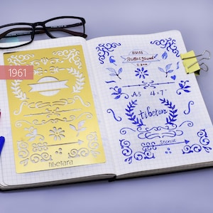 May include: A gold metal stencil with various decorative designs, including swirls, leaves, and arrows. The stencil is labeled "tibetara" and measures 4 x 7 inches. The stencil is on a white notebook page with blue ink markings.