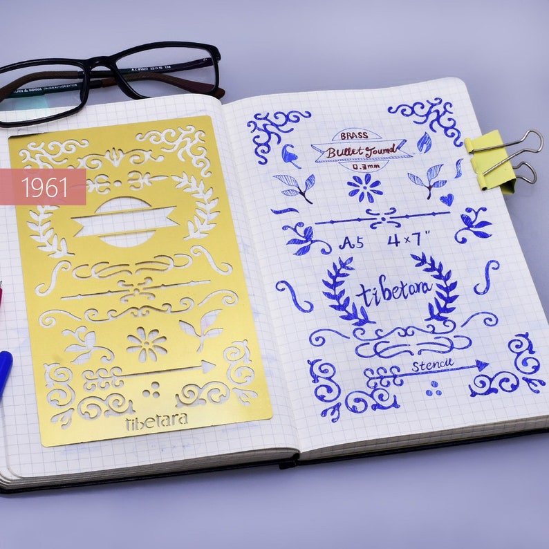 May include: A gold metal stencil with various decorative designs, including swirls, leaves, and arrows. The stencil is labeled "tibetara" and measures 4 x 7 inches. The stencil is on a white notebook page with blue ink markings.