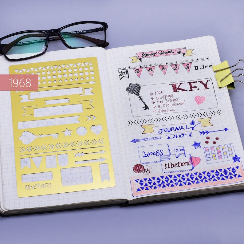 May include: A gold metal stencil with various shapes and designs, including arrows, stars, hearts, and banners. The stencil is labeled "tibetara" and is open on a notebook page with handwritten notes and drawings. The notebook page includes the words "Planner Stencil", "KEY", "JOURNAL", "brass", and "tibetara".