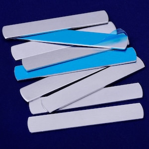 Op de afbeelding: Een stapel zilveren metalen strips met een blauwe kleefrug. De strips zijn ongeveer 1,2 cm breed en 15 cm lang.