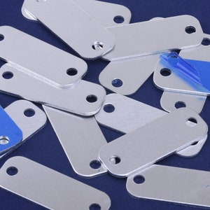 May include: A pile of silver metal rectangular tags with rounded corners and two holes on each tag.