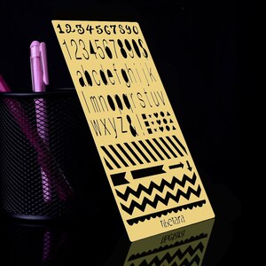 May include: A gold stencil with black numbers, letters, and symbols. The stencil includes the numbers 1-9, the alphabet, and symbols like an ampersand, exclamation point, and stars. The stencil also includes a variety of patterns like stripes, arrows, and zigzags. The stencil is on a black background.