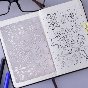 May include: A silver metal stencil featuring a variety of designs, such as butterflies, flowers, and birds. The stencil is positioned on a notebook with a grid pattern, and there are hand-drawn designs on the notebook page.
