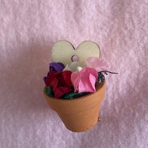 May include: A miniature terracotta flower pot with a wooden heart-shaped accent. The pot is filled with colorful fabric flowers in shades of red, pink, purple, and white. A small gold pin is attached to the pot.