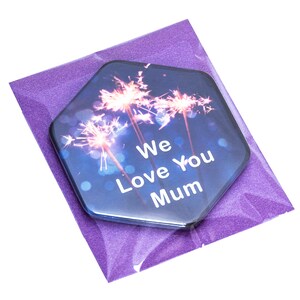 Puede incluir: Un regalo hexagonal con un dise&ntilde;o de fuegos artificiales y el texto "We Love You Mum" se presenta en un sobre morado brillante. El regalo tiene un fondo azul oscuro con luces bokeh.