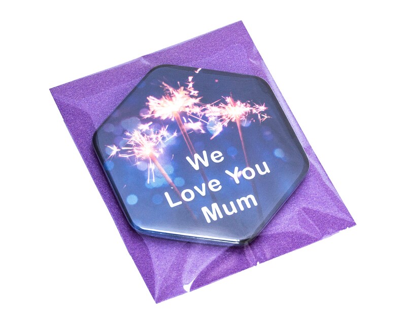 Puede incluir: Un regalo hexagonal con un dise&ntilde;o de fuegos artificiales y el texto "We Love You Mum" se presenta en un sobre morado brillante. El regalo tiene un fondo azul oscuro con luces bokeh.
