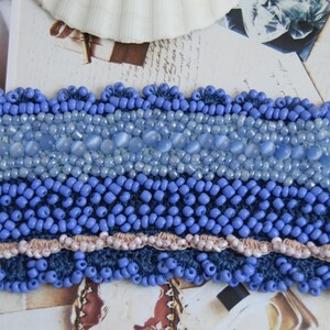 May include: A blue crocheted bracelet with a beaded design. The bracelet features a row of light blue beads, a row of larger, light blue, oval-shaped beads, and a row of smaller, dark blue beads. The bracelet is finished with a scalloped edge.