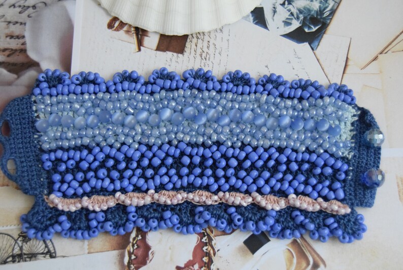 May include: A blue crocheted bracelet with a beaded design. The bracelet features a row of light blue beads, a row of larger, light blue, oval-shaped beads, and a row of smaller, dark blue beads. The bracelet is finished with a scalloped edge.