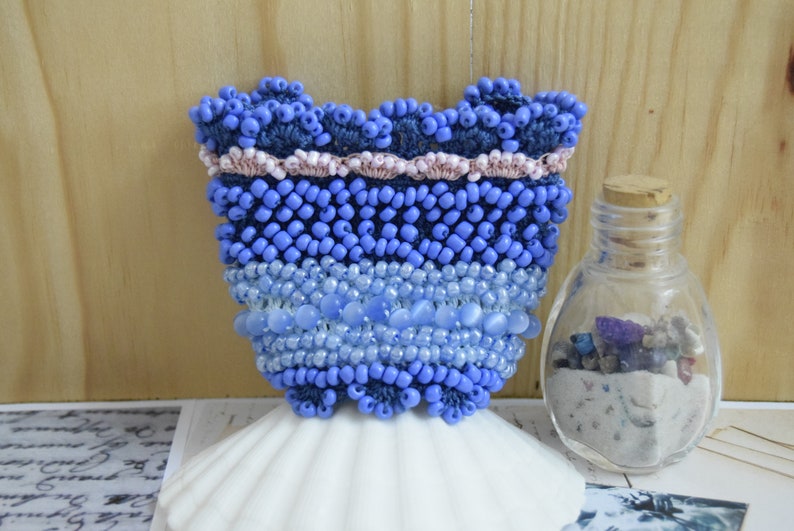 May include: A blue and white beaded purse with a pink trim. The purse is sitting on a white seashell.