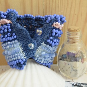 May include: A blue crocheted bracelet with beads and crystals. The bracelet is on a white seashell and a small glass jar with sand and stones.