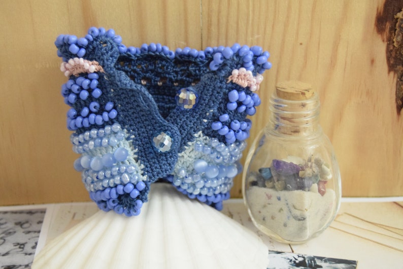 May include: A blue crocheted bracelet with beads and crystals. The bracelet is on a white seashell and a small glass jar with sand and stones.