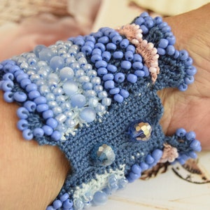 May include: A blue crocheted bracelet with light blue and white beads. The bracelet is decorated with small, iridescent blue and pink beads.