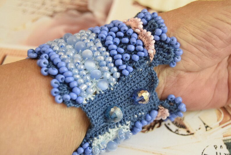 May include: A blue crocheted bracelet with light blue and white beads. The bracelet is decorated with small, iridescent blue and pink beads.