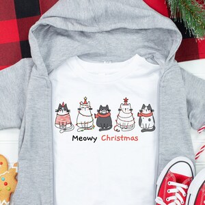 May include: A white hooded sweatshirt with a zipper and a white t-shirt underneath. The t-shirt has a graphic of five cats wearing Christmas hats and the text "Meowy Christmas".