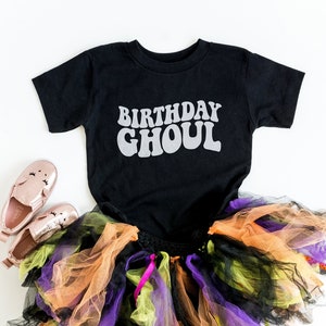 May include: Black t-shirt with the text "Birthday Ghoul" in white, a multi-colored tulle skirt, and pink shoes. Halloween costume for a child.