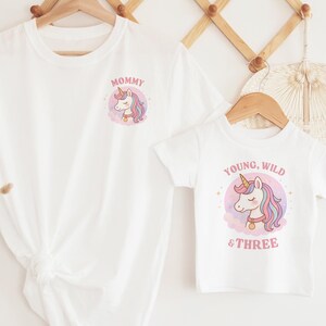 May include: Two white t-shirts, one adult and one child size, hanging on a wooden rack. The adult shirt has the word "Mommy" and a unicorn graphic. The child's shirt says "Young, Wild & Three" with a unicorn.