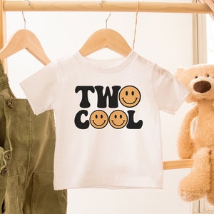 May include: White t-shirt with black text that says "TWO COOL" with three orange smiley faces. The shirt is hanging on a wooden rack with a pair of green overalls and a teddy bear.