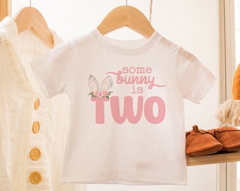 May include: White baby onesie with pink bunny ears and floral accents. The onesie says "some bunny is two" in pink text.