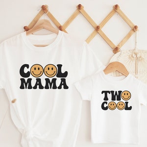 May include: Two white t-shirts with a retro design. The larger shirt says "Cool Mama" with two smiley faces. The smaller shirt says "Two Cool" with two smiley faces.