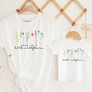 May include: Two white t-shirts with a line of colorful wildflowers printed on them. The larger shirt says "mama of a little wildflower" and the smaller shirt says "little wildflower".