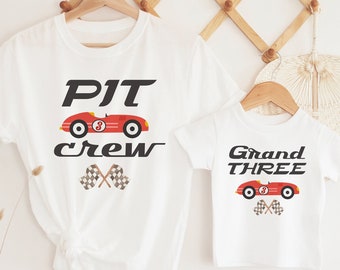 May include: Two white t-shirts with a red race car graphic. The larger shirt says "PIT crew" with checkered flags. The smaller shirt says "Grand THREE" with checkered flags.