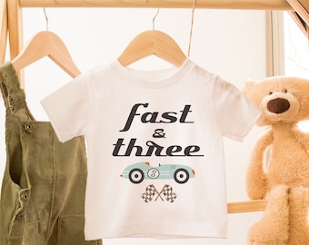 May include: White t-shirt with a blue race car graphic and the text "fast & three". The shirt is hanging on a wooden rack with a pair of green overalls and a teddy bear.