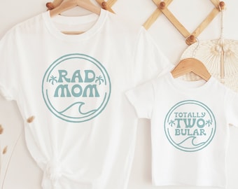 May include: Two white t-shirts with a blue circle design. The larger shirt says "Rad Mom" with palm trees. The smaller shirt says "Totally Two-bular" with palm trees.
