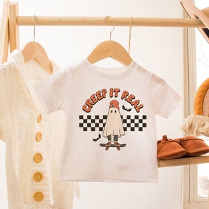 May include: White t-shirt with a graphic of a ghost wearing a red hat and skateboarding. The text "Creep it Real" is printed above the ghost. The t-shirt is hanging on a wooden rack.