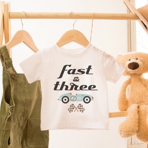 Könnte beinhalten: Weißes T-Shirt mit einem blauen Rennwagen-Motiv und dem Text "fast & three". Das Shirt hängt an einem Holzgestell mit einer grünen Latzhose und einem Teddybär.