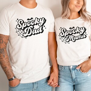 May include: A couple wearing white t-shirts with black text that says "Spooky Dad" and "Spooky Mama" with a spiderweb and bat graphic.