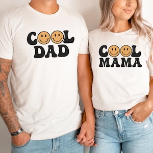 May include: Two white t-shirts with black text and smiley faces. The text on the first shirt says "COOL DAD" and the text on the second shirt says "COOL MAMA".