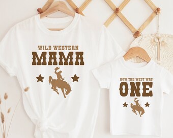 May include: Two white t-shirts with brown text and a cowboy riding a horse graphic. The larger shirt says "Wild Western Mama" and the smaller shirt says "How the West Was One".