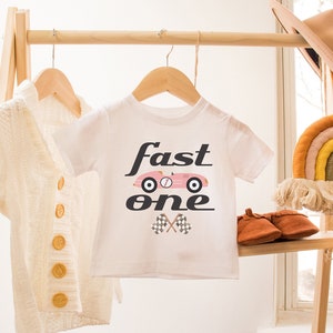 May include: White baby t-shirt with a pink race car graphic and the text "fast one" in black. The shirt is hanging on a wooden rack with a white cardigan and a rainbow wall hanging.
