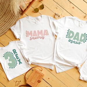 May include: Four white t-shirts with green and pink dinosaur themed text. The shirts say 'Mama saurus', 'Dad osaur', 'Sister saurus', and 'Rex 3'. The shirts are laid out on a wooden surface.