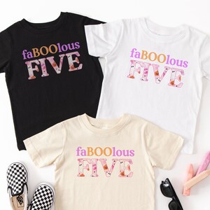 May include: Three children's t-shirts in black, white, and cream, each printed with the words "faBOOlous FIVE" in purple and orange. A pair of black and white chequered slip-on shoes, sunglasses, and a decorative item are also visible.