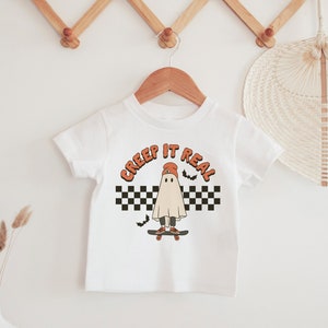 May include: White t-shirt with a ghost wearing a red hat and skateboarding. The text "CREEP IT REAL" is above the ghost. The ghost is on a checkered background with bats flying around it.
