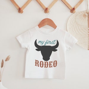 May include: White baby t-shirt with a black bull silhouette and the text "my first rodeo" in brown and teal.