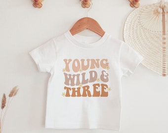 May include: White t-shirt with the text "YOUNG WILD & THREE" in brown, orange, and pink lettering with small yellow flower accents.