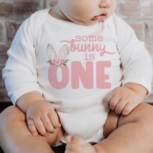 May include: A white long-sleeved onesie with pink bunny ears and a pink floral crown. The onesie has the text "some bunny is one" in pink.
