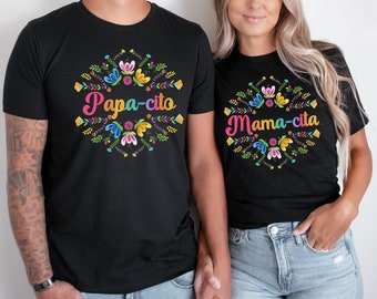 May include: Black t-shirts with floral designs and text. One shirt says "Papa-cito" in colorful letters, the other says "Mama-cita". The shirts have a round floral wreath design around the text.