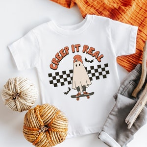 May include: White t-shirt with a graphic of a ghost wearing a red beanie and skateboarding. The text "CREEP IT REAL" is above the ghost.