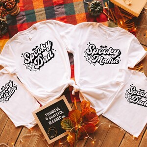 May include: Four white t-shirts with black text and spiderwebs. The shirts say "Spooky Dad", "Spooky Mama", "Spooky Babe", and "Spooky Dude".