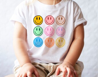 May include: A white t-shirt with a colorful smiley face design. The shirt features nine different colored smiley faces arranged in a 3x3 grid.