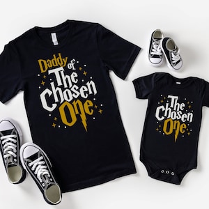 May include: Two black t-shirts with gold lettering. The larger shirt says "Daddy of The Chosen One" and the smaller shirt says "The Chosen One".