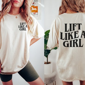 K&ouml;nnte beinhalten: Ein cremefarbenes T-Shirt mit dem Text "Lift Like A Girl" in Schwarz auf Vorder- und R&uuml;ckseite.