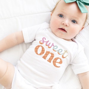 May include: A white onesie with the words "sweet one" in colorful letters with a waffle cone pattern. The onesie is worn by a baby with a green bow in their hair.