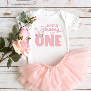 May include: A white onesie with pink bunny ears and flowers, and the text "some bunny is one". The onesie is paired with a pink tulle tutu.