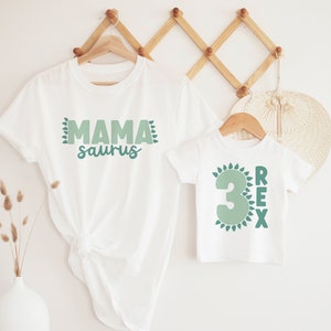 May include: Two white t-shirts with green dinosaur-themed text. The larger shirt says "Mama saurus" and the smaller shirt says "3 Rex" with a green number 3.