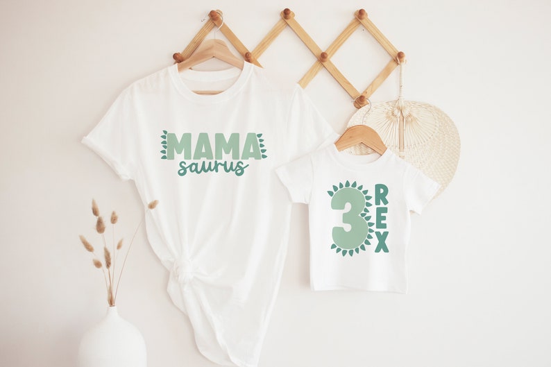 May include: Two white t-shirts with green dinosaur-themed text. The larger shirt says "Mama saurus" and the smaller shirt says "3 Rex" with a green number 3.