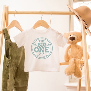 May include: A white t-shirt with a blue circle graphic that says "THE BIG ONE" with palm trees and a wave. The shirt is hanging on a wooden rack with a pair of green overalls and a teddy bear.